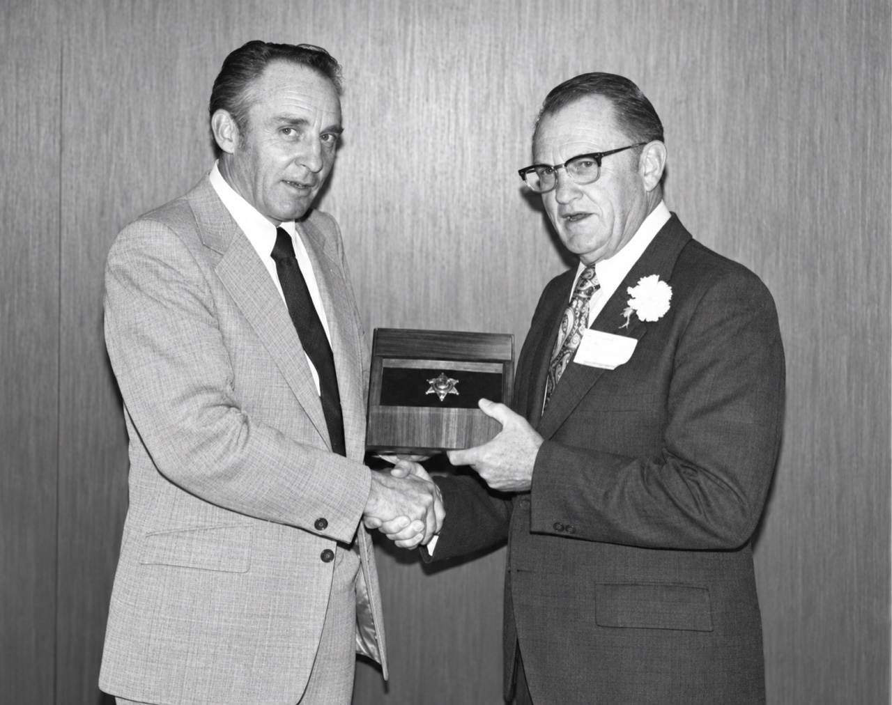 Two men in suits shaking hands, one presenting a wooden case with a star badge mounted in it