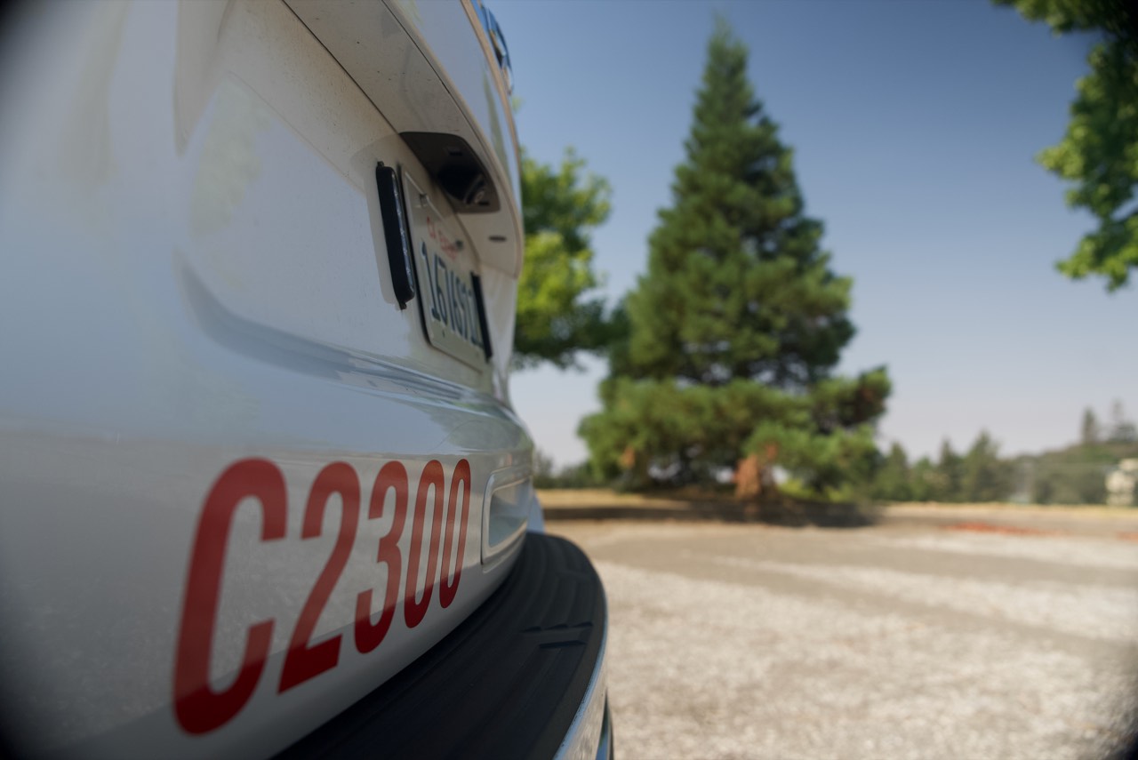 Close view along the front of a white CAL FIRE vehicle lettered C2300, with Chief Waggoner's redwood standing on the far side of the yard